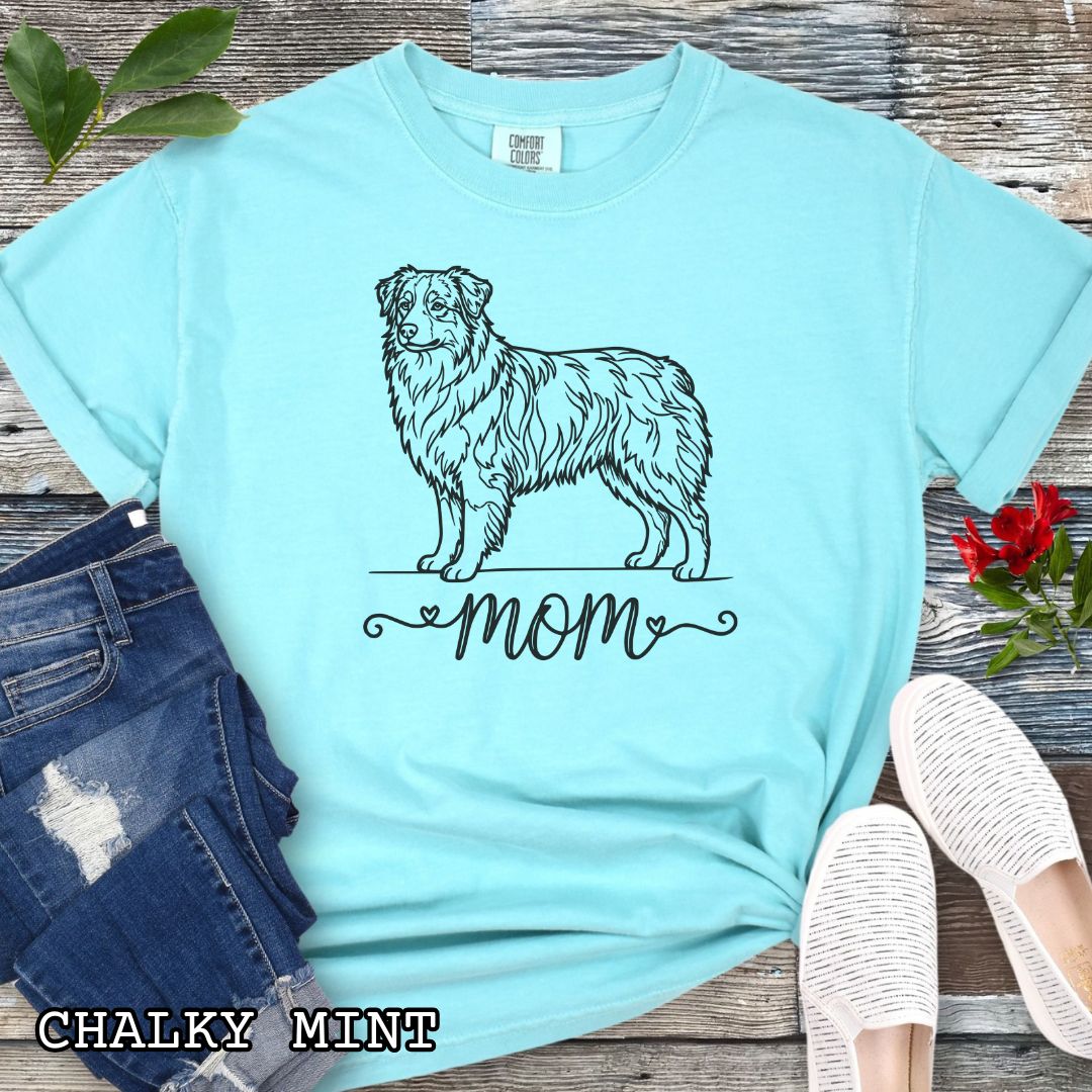 Mint Comfort Colors t-shirt with a line art Australian Shepherd dog illustration and 'mom' text in feminine script beneath, shown on a wooden surface with denim jeans and a flower.