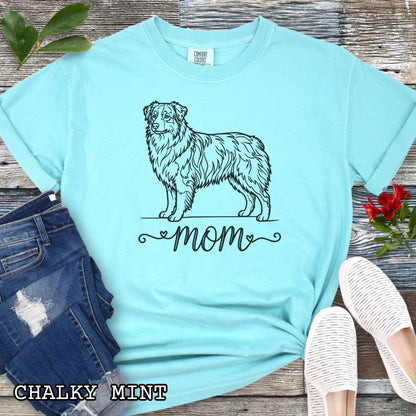 Mint Comfort Colors t-shirt with a line art Australian Shepherd dog illustration and 'mom' text in feminine script beneath, shown on a wooden surface with denim jeans and a flower.