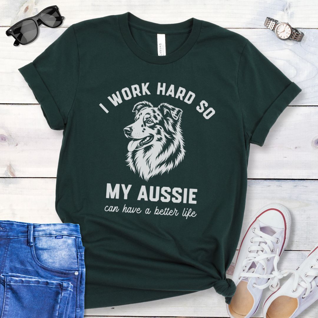 Dark green unisex t-shirt for Australian Shepherd lovers with breed head study graphic and humorous text 'I work hard so my Aussie can have a better life', shown on a wooden surface with jeans and shoes. 