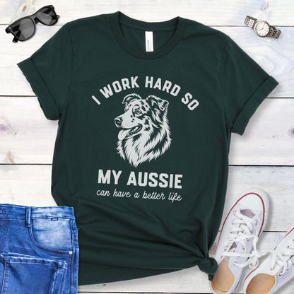 Dark green unisex t-shirt for Australian Shepherd lovers with breed head study graphic and humorous text 'I work hard so my Aussie can have a better life', shown on a wooden surface with jeans and shoes. 