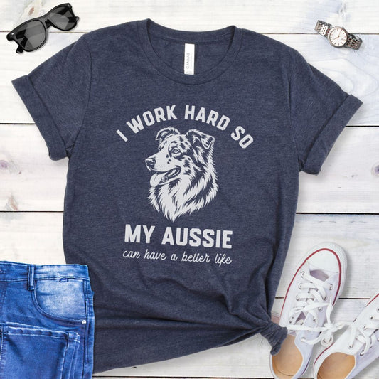 Heather navy blue unisex t-shirt for Australian Shepherd lovers with breed head study graphic and humorous text 'I work hard so my Aussie can have a better life', shown on a wooden surface with jeans and shoes. 