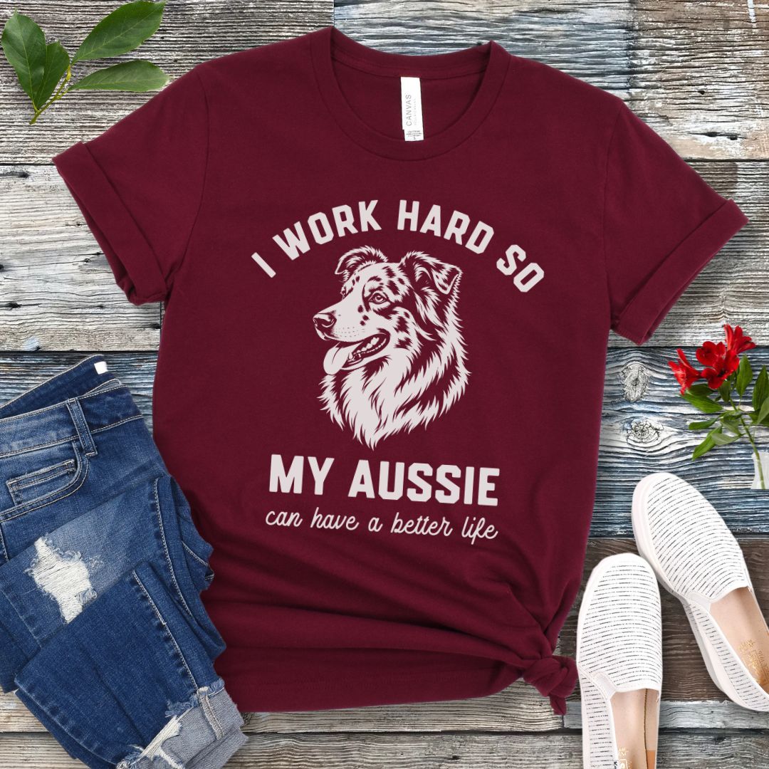 Maroon unisex t-shirt for Australian Shepherd lovers with breed head study graphic and humorous text 'I work hard so my Aussie can have a better life', shown on a wooden surface with jeans and shoes. 