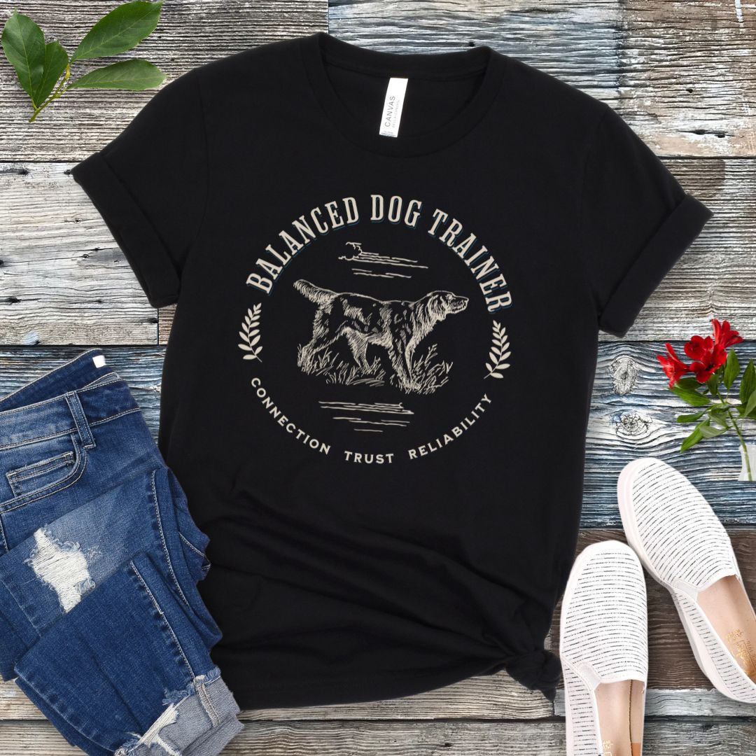 A black vintage-style t-shirt with the text 'Balanced Dog Trainer' and a graphic of a dog, presented on a wooden surface with a pair of jeans and white shoes in the background.