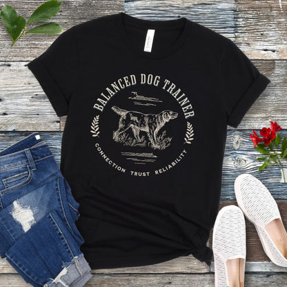 A black vintage-style t-shirt with the text 'Balanced Dog Trainer' and a graphic of a dog, presented on a wooden surface with a pair of jeans and white shoes in the background.