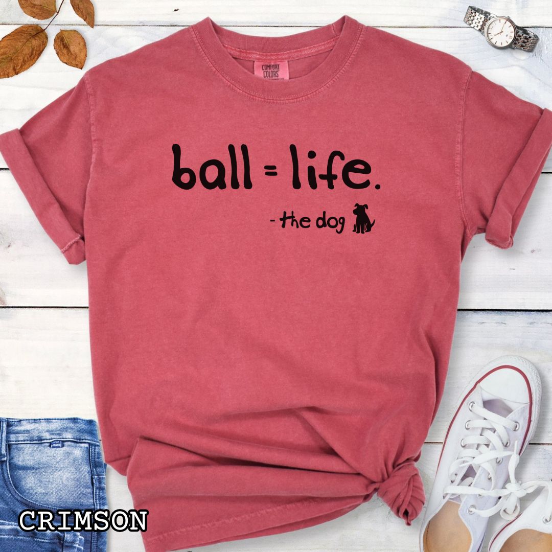 Red Comfort Colors t-shirt for dog lovers with humorous text 'ball = life. - the dog' in playful hand-drawn style lettering, accompanied by a dog graphic, shown on a wooden surface with jeans and flowers.