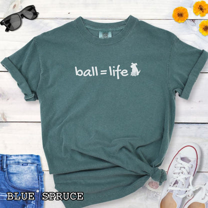 Green Comfort Colors t-shirt for dog lovers with humorous text 'ball = life' in playful hand-drawn style lettering, accompanied by a dog graphic, shown on a wooden surface with jeans and flowers.