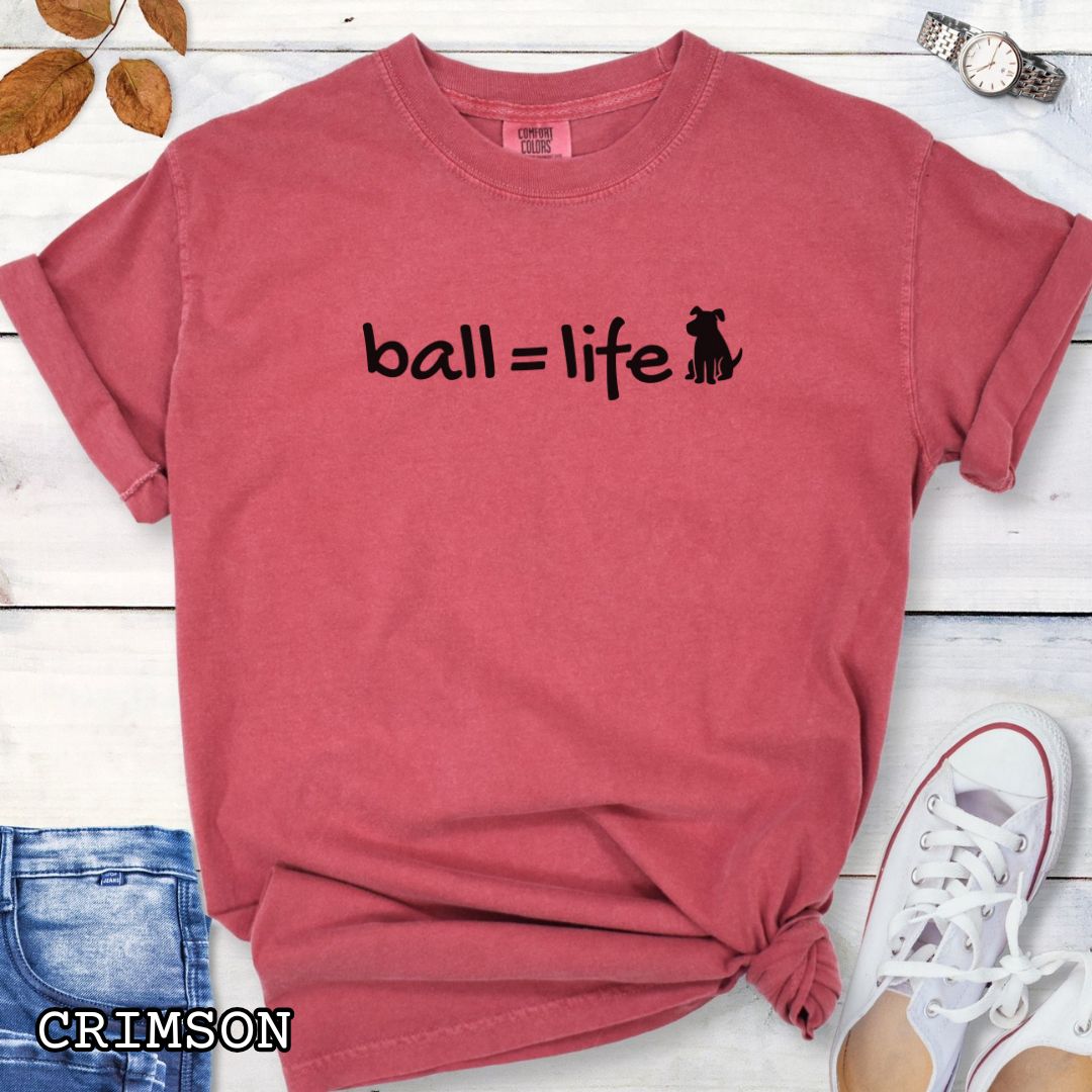 Red Comfort Colors t-shirt for dog lovers with humorous text 'ball = life' in playful hand-drawn style lettering, accompanied by a dog graphic, shown on a wooden surface with jeans and flowers.