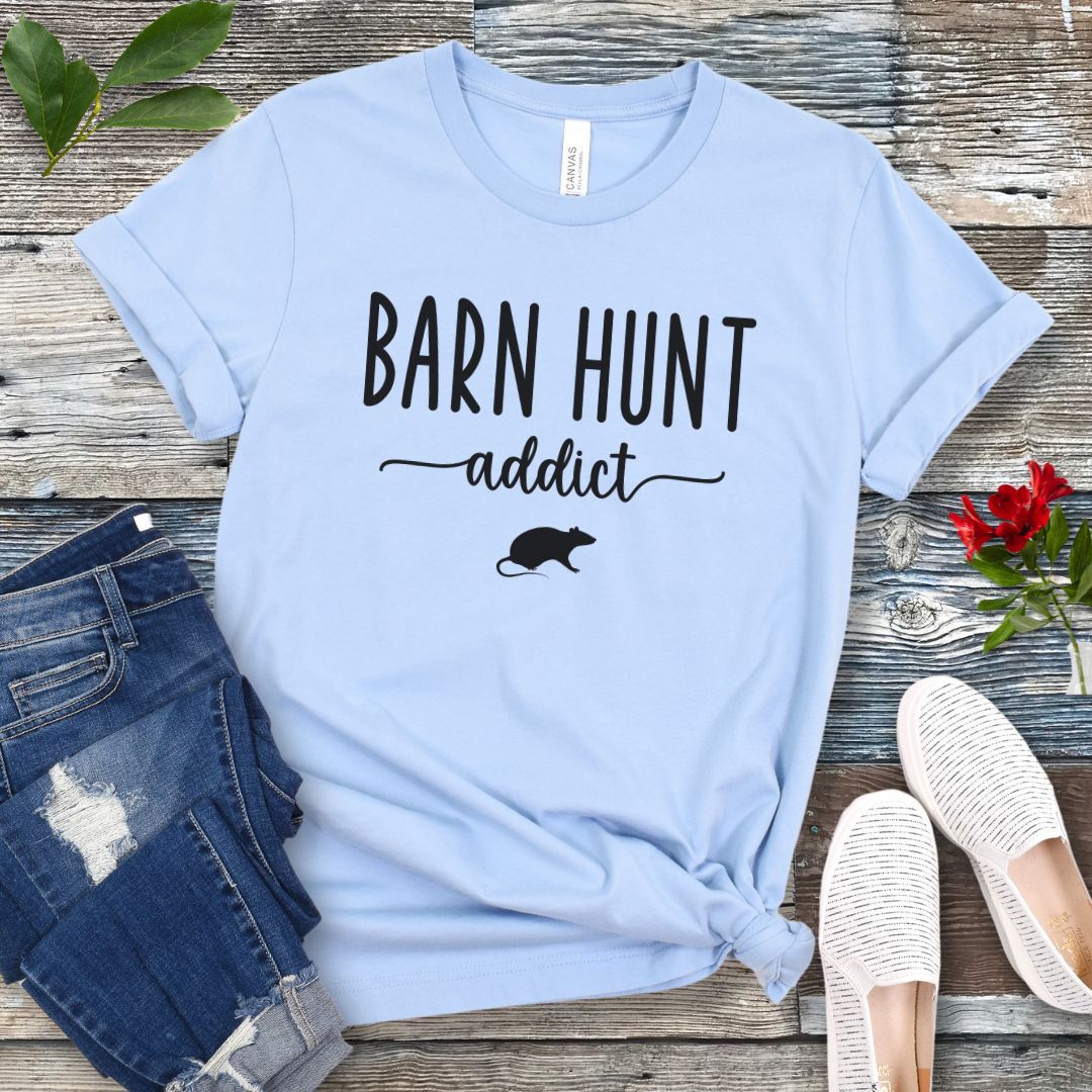 Light blue dog sports t-shirt with 'Barn Hunt Addict' text and a rat graphic on a wooden surface with jeans and shoes.