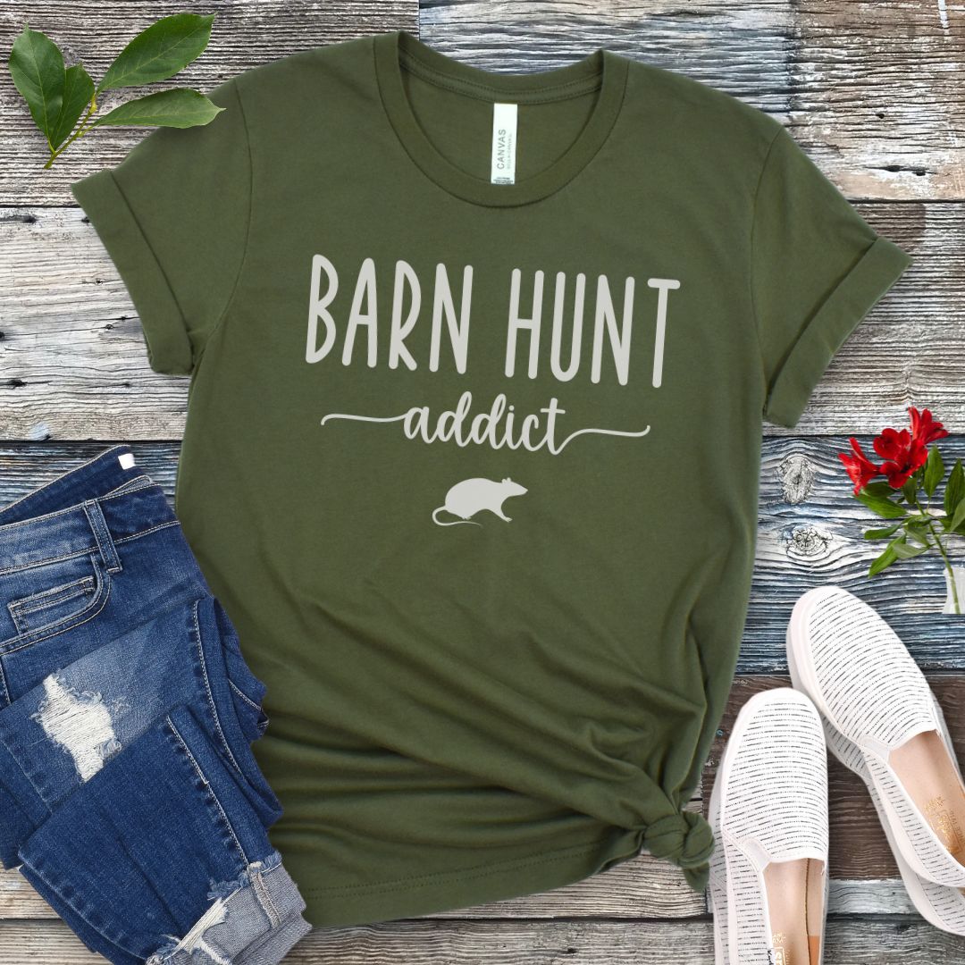 Military green dog sports t-shirt with 'Barn Hunt Addict' text and a rat graphic on a wooden surface with jeans and shoes.