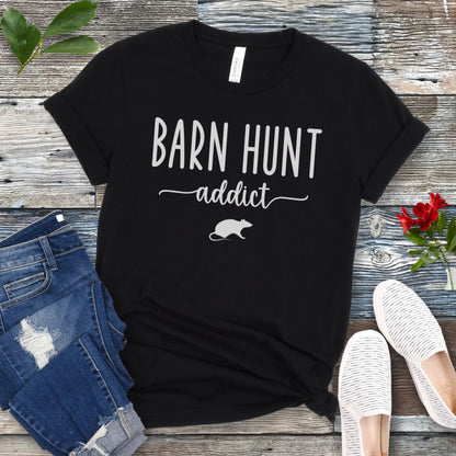 Black dog sports t-shirt with 'Barn Hunt Addict' text and a rat graphic on a wooden surface with jeans and shoes.