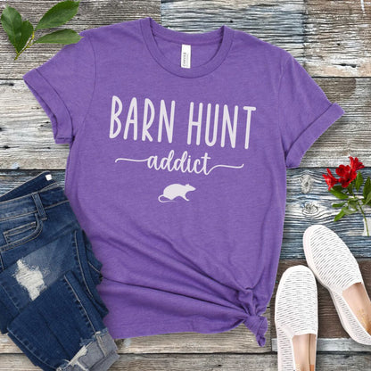 Purple dog sports t-shirt with 'Barn Hunt Addict' text and a rat graphic on a wooden surface with jeans and shoes.