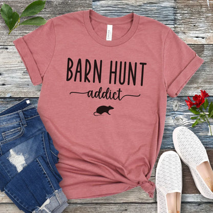 Mauve dog sports t-shirt with 'Barn Hunt Addict' text and a rat graphic on a wooden surface with jeans and shoes.