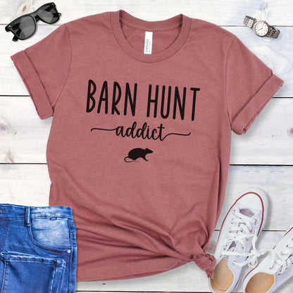 Mauve dog sports t-shirt with 'Barn Hunt Addict' text and a rat graphic on a wooden surface with jeans and shoes.
