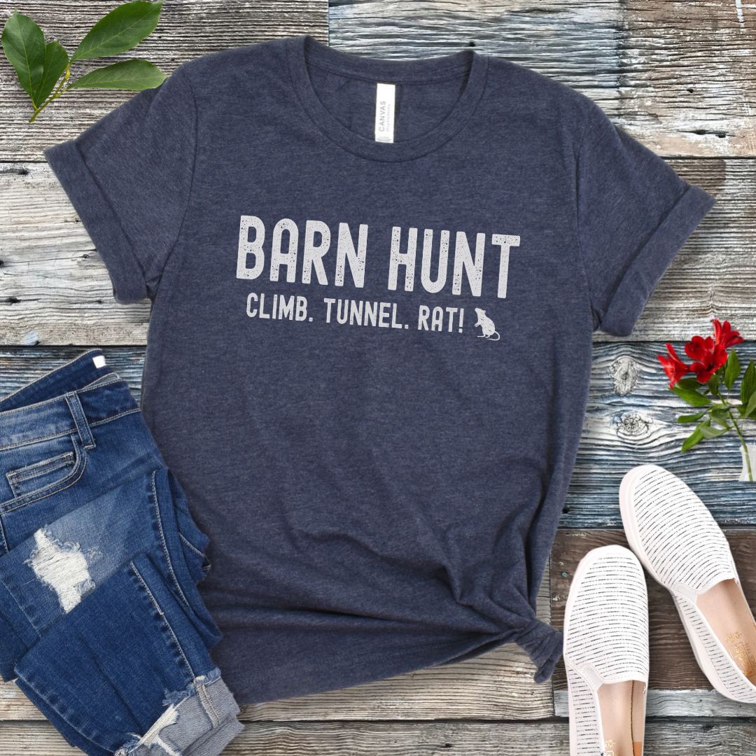 Heather navy blue dog sports t-shirt with 'Barn Hunt - Climb, Tunnel, Rat!' text on a wooden surface with jeans and shoes.