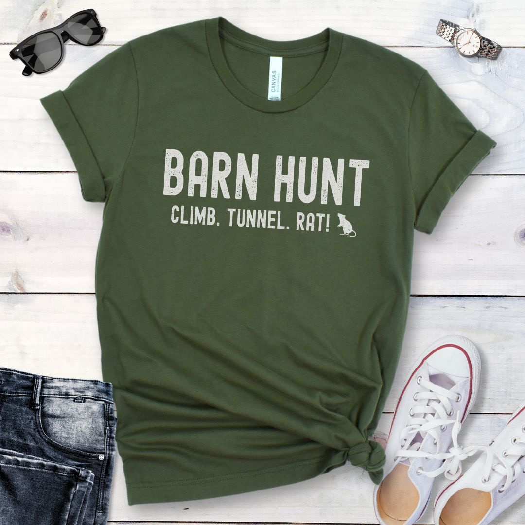Military green dog sports t-shirt with 'Barn Hunt - Climb, Tunnel, Rat!' text on a wooden surface with jeans and shoes.