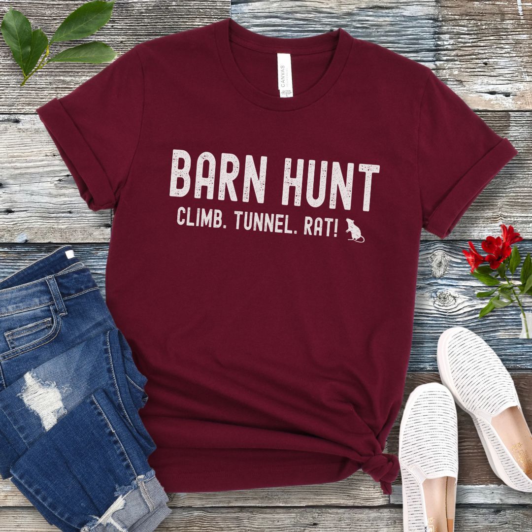 Maroon dog sports t-shirt with 'Barn Hunt - Climb, Tunnel, Rat!' text on a wooden surface with jeans and shoes.