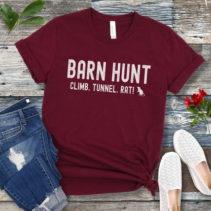 Maroon dog sports t-shirt with 'Barn Hunt - Climb, Tunnel, Rat!' text on a wooden surface with jeans and shoes.