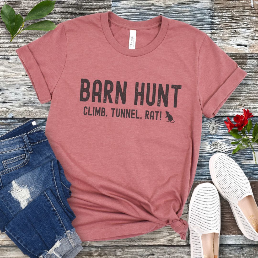 Mauve dog sports t-shirt with 'Barn Hunt - Climb, Tunnel, Rat!' text on a wooden surface with jeans and shoes.