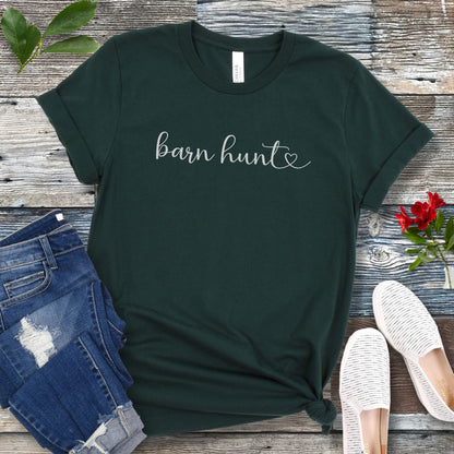 Forest green dog sports t-shirt with 'barn hunt' text in handwritten script and heart, presented on a wooden surface with jeans and shoes.