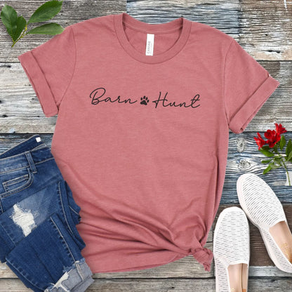 Mauve dog sports t-shirt with 'Barn Hunt' script text with small pawprint, presented on a wooden surface with jeans and shoes.