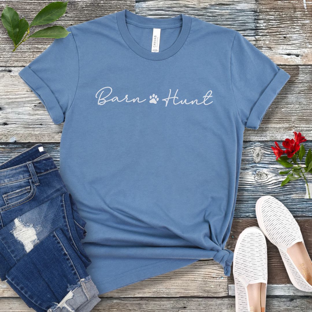Blue dog sports t-shirt with 'Barn Hunt' script text with small pawprint, presented on a wooden surface with jeans and shoes.