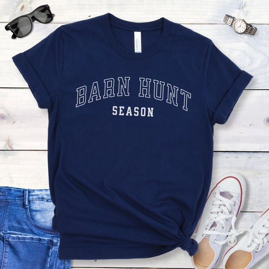 Navy blue dog sports t-shirt with 'Barn Hunt Season' in college-style text, presented on a wooden surface with jeans and shoes.