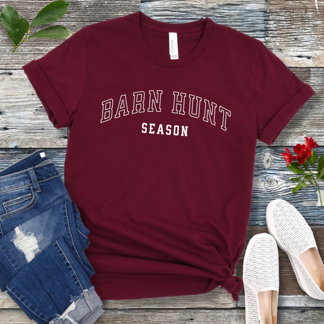Maroon dog sports t-shirt with 'Barn Hunt Season' in college-style text, presented on a wooden surface with jeans and shoes.