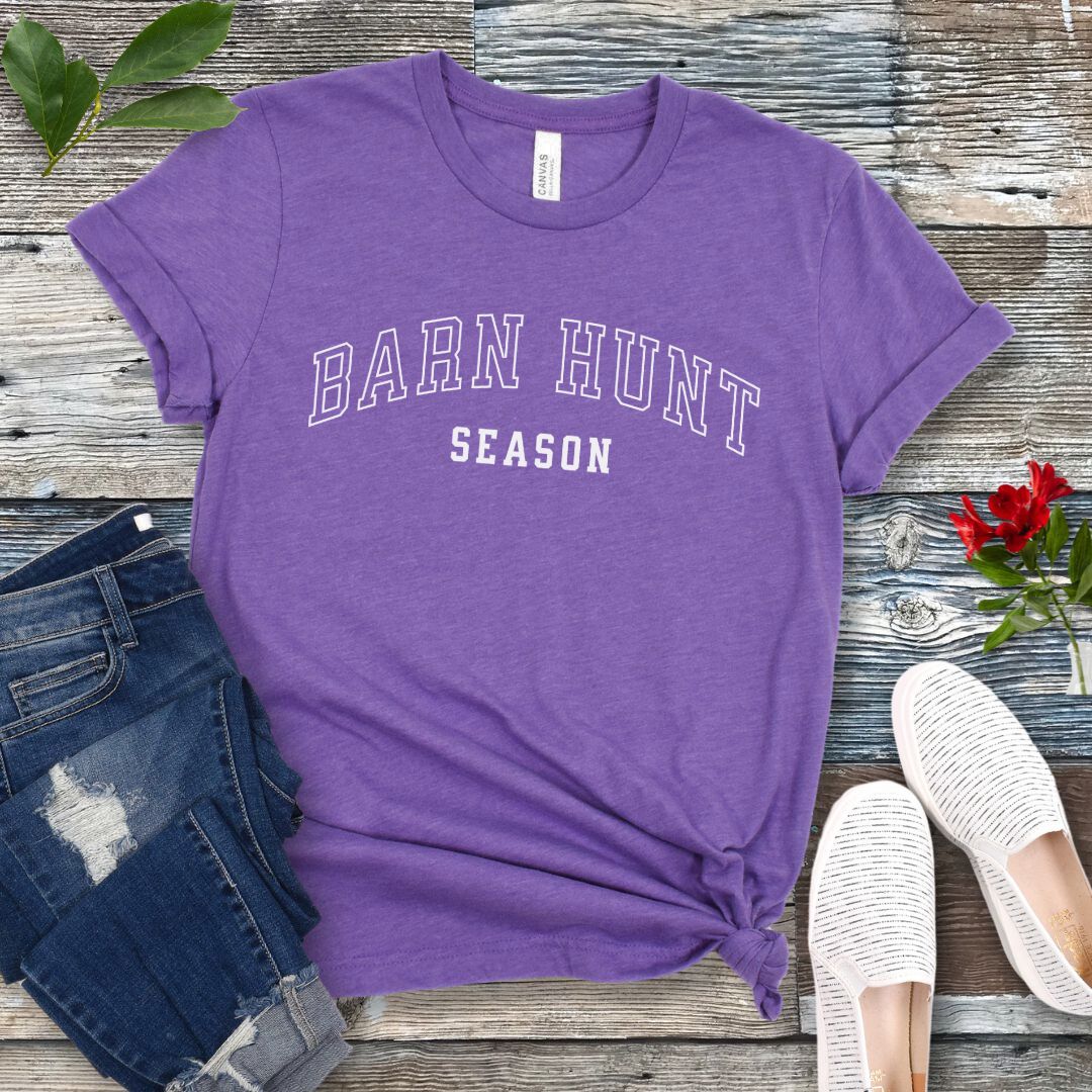 Purple dog sports t-shirt with 'Barn Hunt Season' in college-style text, presented on a wooden surface with jeans and shoes.