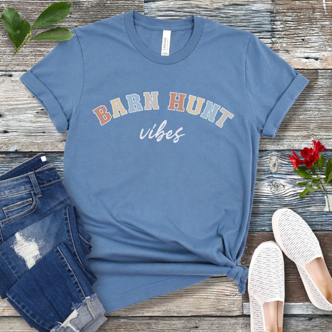 Blue dog sports t-shirt with 'Barn Hunt Vibes' text on a wooden surface with jeans and shoes.