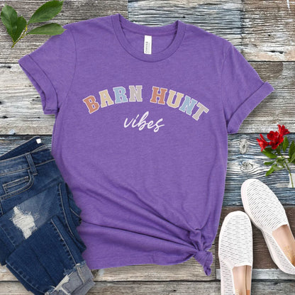 Purple dog sports t-shirt with 'Barn Hunt Vibes' text on a wooden surface with jeans and shoes.