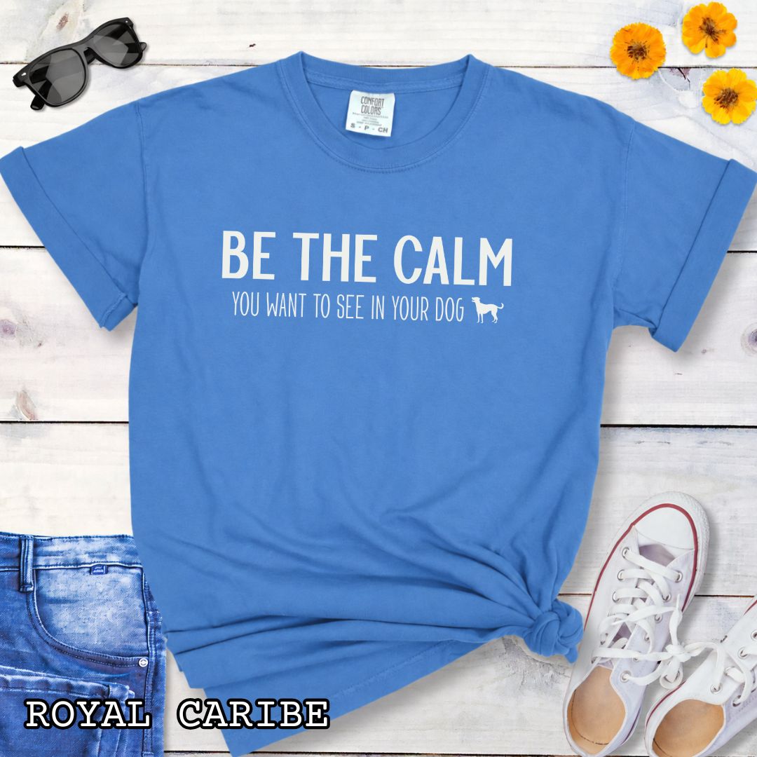 Royal blue Comfort Colors t-shirt with text 'BE THE CALM YOU WANT TO SEE IN YOUR DOG' on a wooden surface with jeans and a plant.