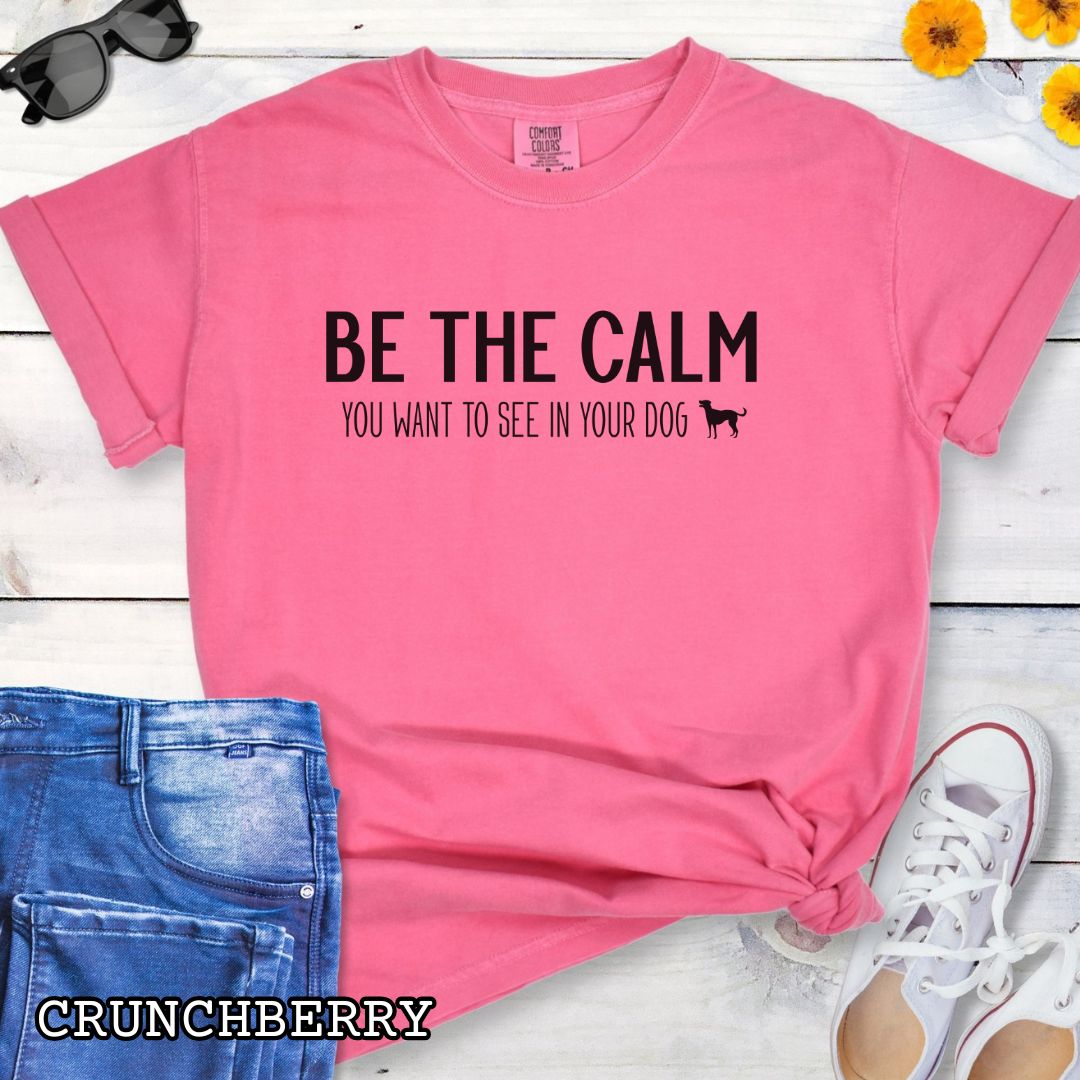 Bright Pink color Comfort Colors t-shirt with text 'BE THE CALM YOU WANT TO SEE IN YOUR DOG' on a wooden surface with jeans and a plant.