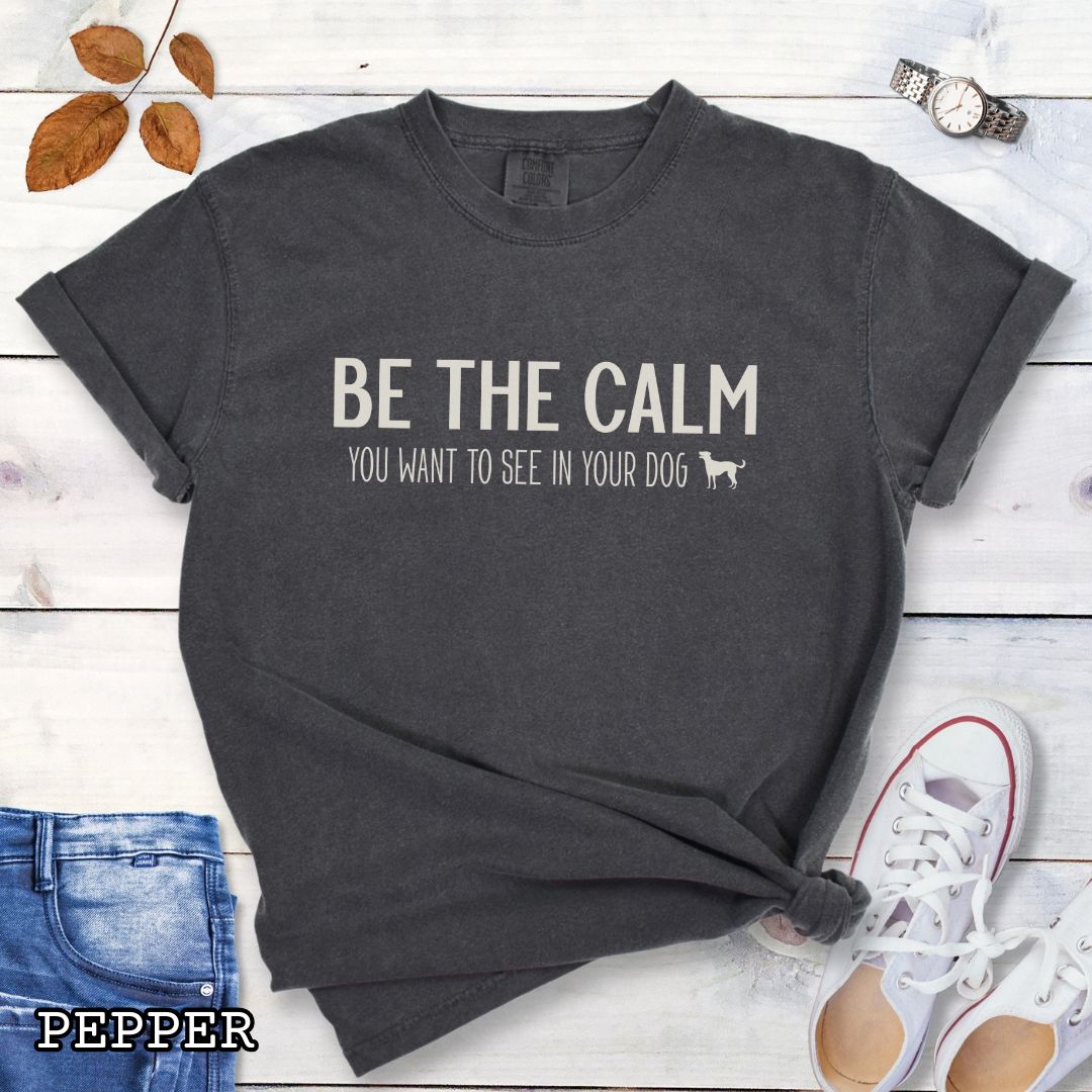 Dark grey (Pepper) Comfort Colors t-shirt with text 'BE THE CALM YOU WANT TO SEE IN YOUR DOG' on a wooden surface with jeans and a plant.