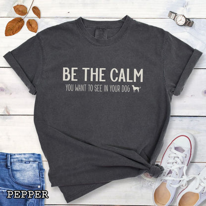 Dark grey (Pepper) Comfort Colors t-shirt with text 'BE THE CALM YOU WANT TO SEE IN YOUR DOG' on a wooden surface with jeans and a plant.