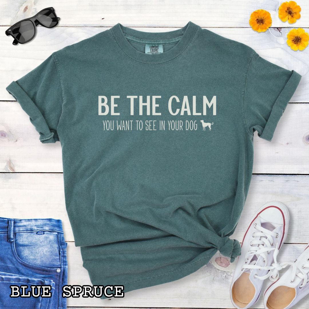 Green (Blue Spruce) Comfort Colors t-shirt with text 'BE THE CALM YOU WANT TO SEE IN YOUR DOG' on a wooden surface with jeans and a plant.