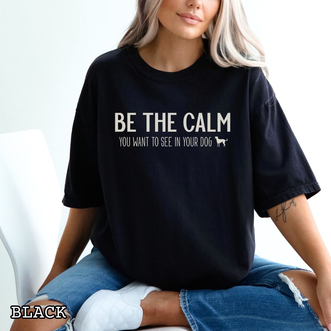 Person wearing a black Comfort Colors -shirt with 'Be The Calm You Want to See In Your Dog' text sitting on a white couch.