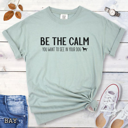 Light green (Bay) Comfort Colors t-shirt with text 'BE THE CALM YOU WANT TO SEE IN YOUR DOG' on a wooden surface with jeans and a plant.