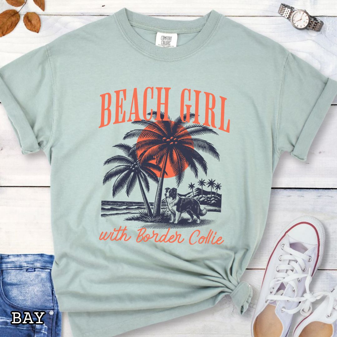 Light green Comfort Colors t-shirt for Border Collie lovers with graphic of a Border Collie standing on a tropical beach, and the text 'Beach Girl With Border Collie' in retro lettering, shown on a wooden surface with accessories.