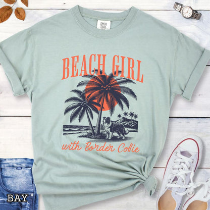 Light green Comfort Colors t-shirt for Border Collie lovers with graphic of a Border Collie standing on a tropical beach, and the text 'Beach Girl With Border Collie' in retro lettering, shown on a wooden surface with accessories.