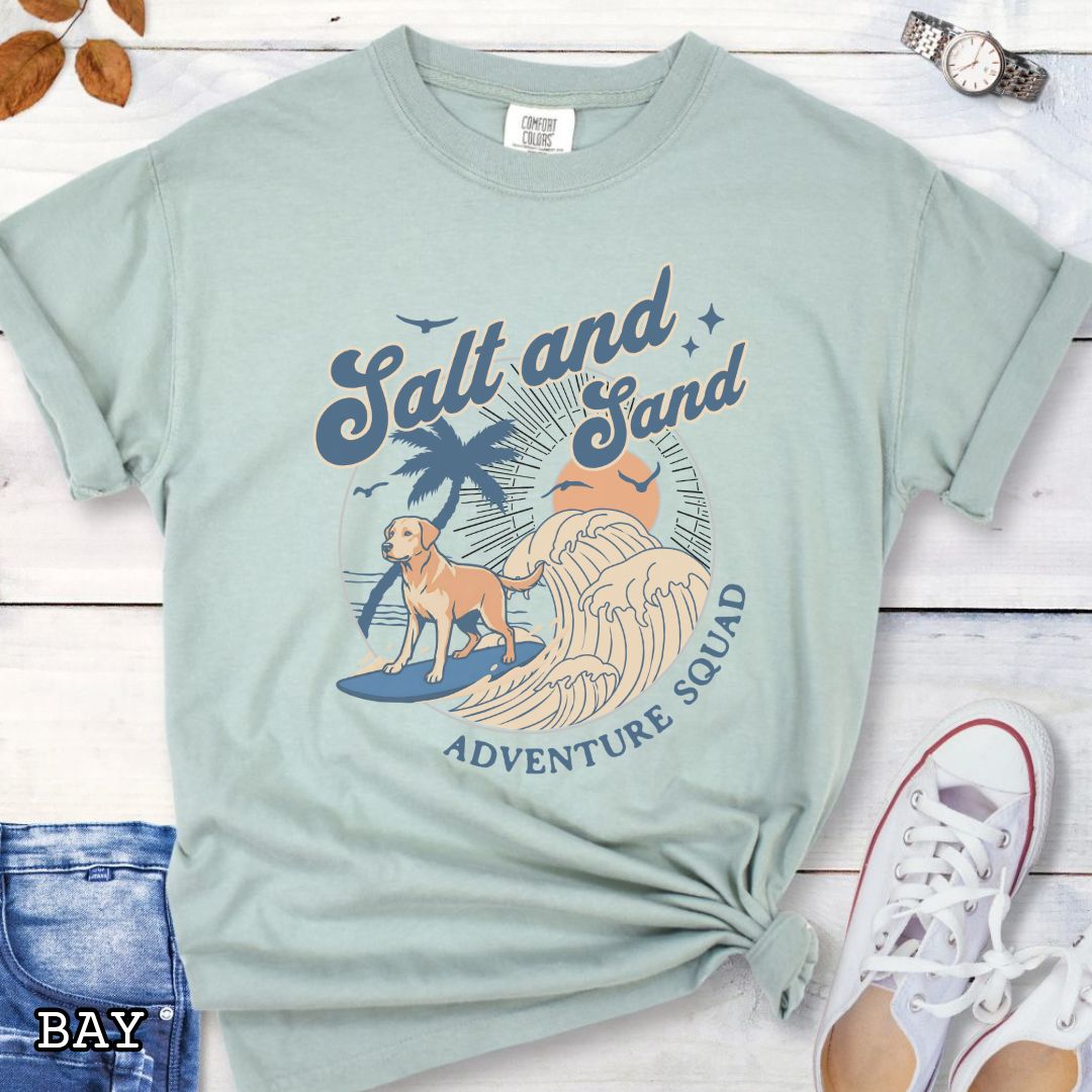 Bay Comfort Colors t-shirt for Labrador Retriever lovers with beach-themed graphic of a surfing Labrador, waves and sunset, and the text 'Salt and Sand Adventure Squad' in retro lettering, shown on a wooden surface with accessories.