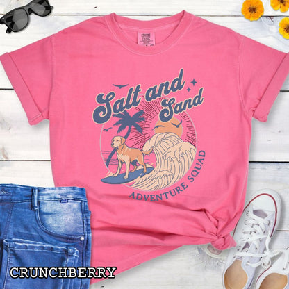 Bright pink Comfort Colors t-shirt for Labrador Retriever lovers with beach-themed graphic of a surfing Labrador, waves and sunset, and the text 'Salt and Sand Adventure Squad' in retro lettering, shown on a wooden surface with accessories.