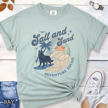 Light green Comfort Colors t-shirt for Belgian Sheepdog lovers with beach-themed graphic of a surfing Belgian Sheepdog, waves and sunset, and the text 'Salt and Sand Adventure Squad' in retro lettering, shown on a wooden surface with accessories.