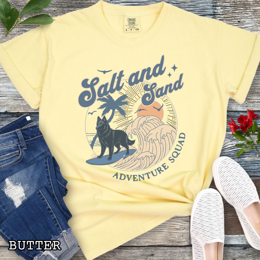 Yellow Comfort Colors t-shirt for Belgian Sheepdog lovers with beach-themed graphic of a surfing Belgian Sheepdog, waves and sunset, and the text 'Salt and Sand Adventure Squad' in retro lettering, shown on a wooden surface with accessories.
