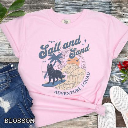 Pink Comfort Colors t-shirt for Belgian Sheepdog lovers with beach-themed graphic of a surfing Belgian Sheepdog, waves and sunset, and the text 'Salt and Sand Adventure Squad' in retro lettering, shown on a wooden surface with accessories.