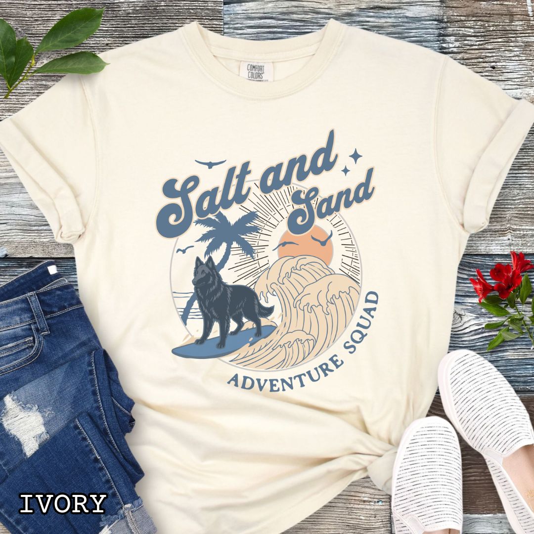 Beige Comfort Colors t-shirt for Belgian Sheepdog lovers with beach-themed graphic of a surfing Belgian Sheepdog, waves and sunset, and the text 'Salt and Sand Adventure Squad' in retro lettering, shown on a wooden surface with accessories.