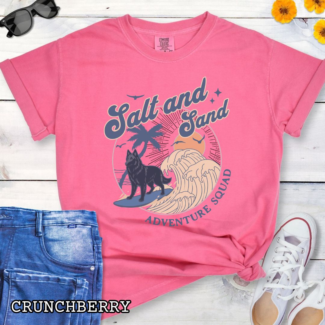 Bright pink Comfort Colors t-shirt for Belgian Sheepdog lovers with beach-themed graphic of a surfing Belgian Sheepdog, waves and sunset, and the text 'Salt and Sand Adventure Squad' in retro lettering, shown on a wooden surface with accessories.