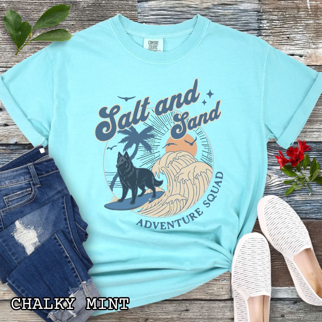 Mint Comfort Colors t-shirt for Belgian Sheepdog lovers with beach-themed graphic of a surfing Belgian Sheepdog, waves and sunset, and the text 'Salt and Sand Adventure Squad' in retro lettering, shown on a wooden surface with accessories.