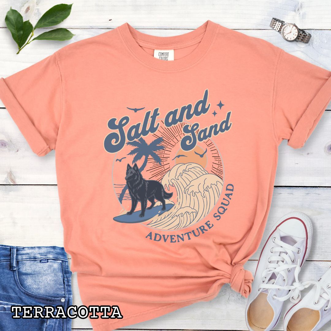 Orange Comfort Colors t-shirt for Belgian Sheepdog lovers with beach-themed graphic of a surfing Belgian Sheepdog, waves and sunset, and the text 'Salt and Sand Adventure Squad' in retro lettering, shown on a wooden surface with accessories.