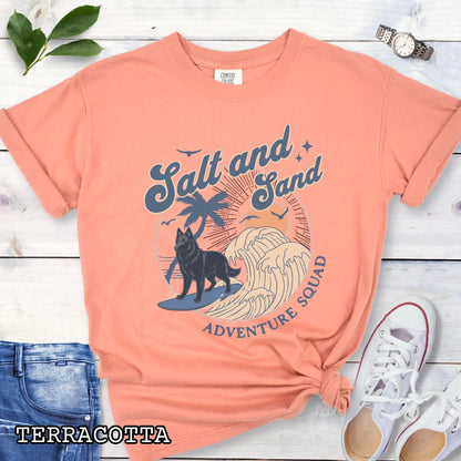 Orange Comfort Colors t-shirt for Belgian Sheepdog lovers with beach-themed graphic of a surfing Belgian Sheepdog, waves and sunset, and the text 'Salt and Sand Adventure Squad' in retro lettering, shown on a wooden surface with accessories.