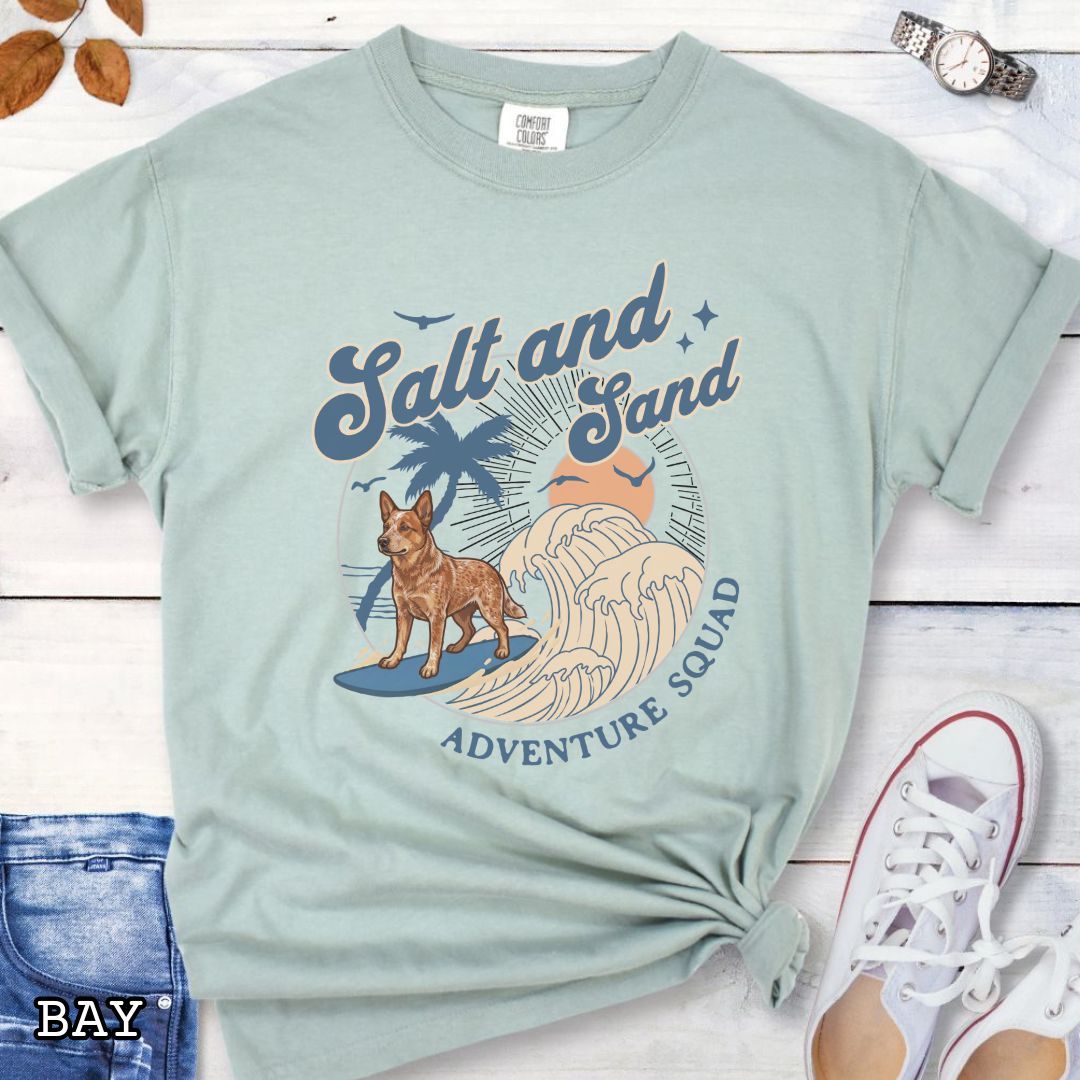 Bay Comfort Colors t-shirt for Australian Cattle Dog lovers with beach-themed graphic of a surfing Red Heeler, waves and sunset, and the text 'Salt and Sand Adventure Squad' in retro lettering, shown on a wooden surface with accessories.